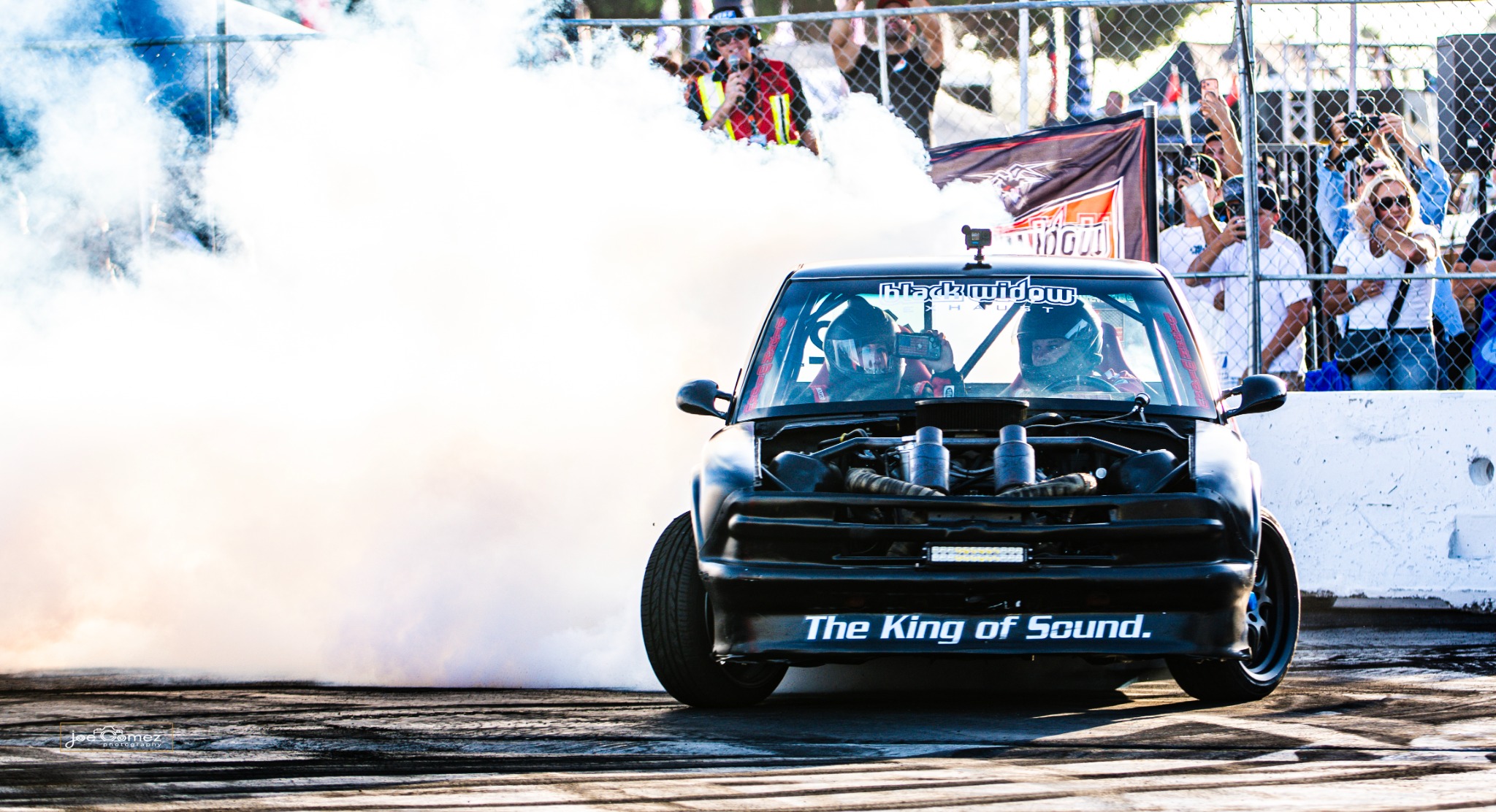 A black off-road vehicle with helmeted occupants surrounded by burnout smoke and spectators