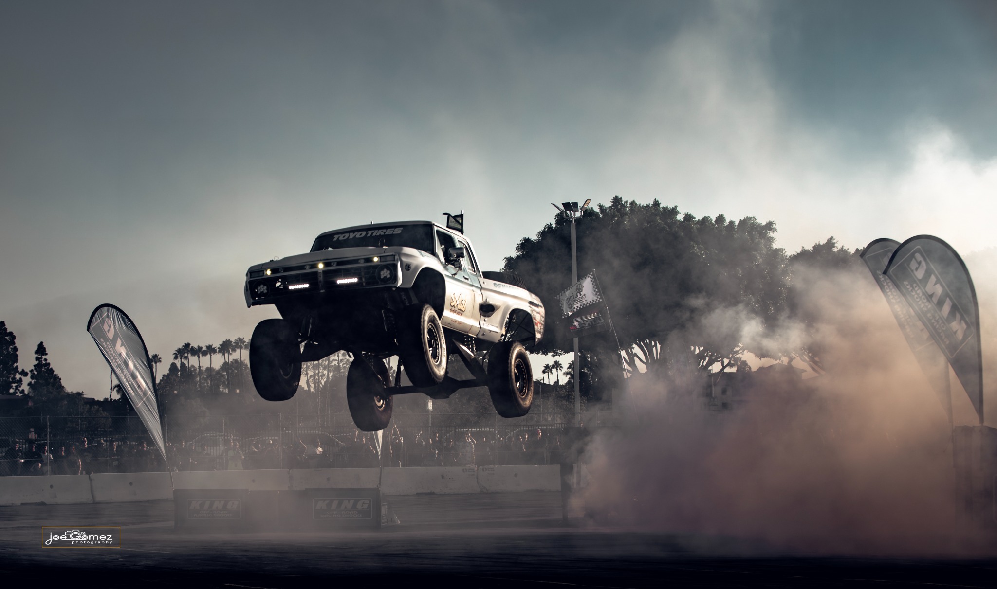 A silver off-road truck hangs above the arena surface beside a cloud of burnout smoke