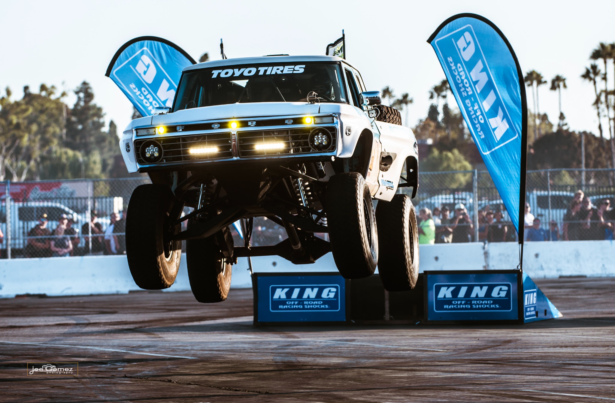 A white off-road truck airborne above the arena, its headlights illuminated