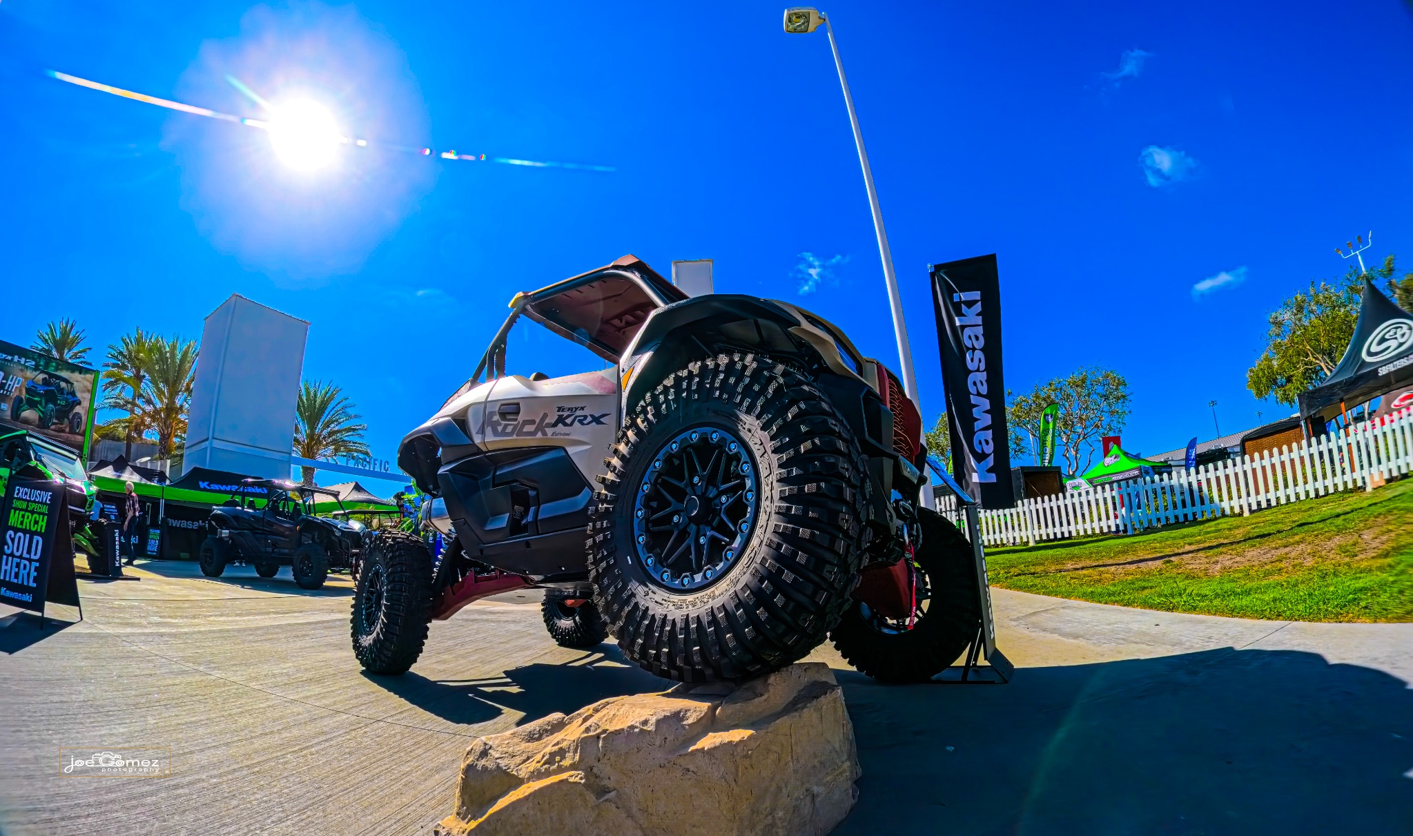A Kawasaki side-by-side sits with one front tire on a display rock beneath the bright afternoon sun