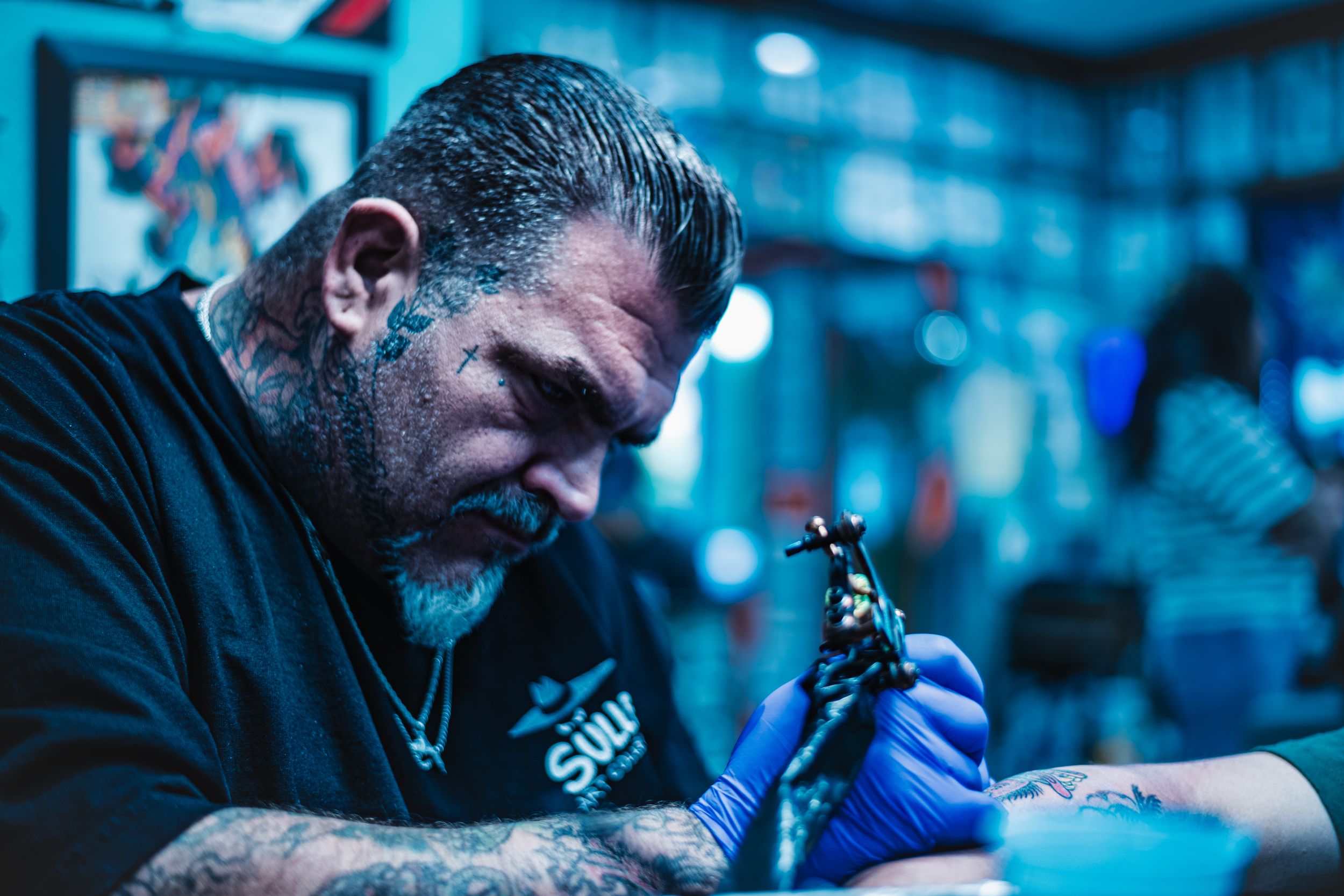 Blue-toned behind-the-scenes portrait of an older tattoo artist working with focused precision in a Los Angeles studio.