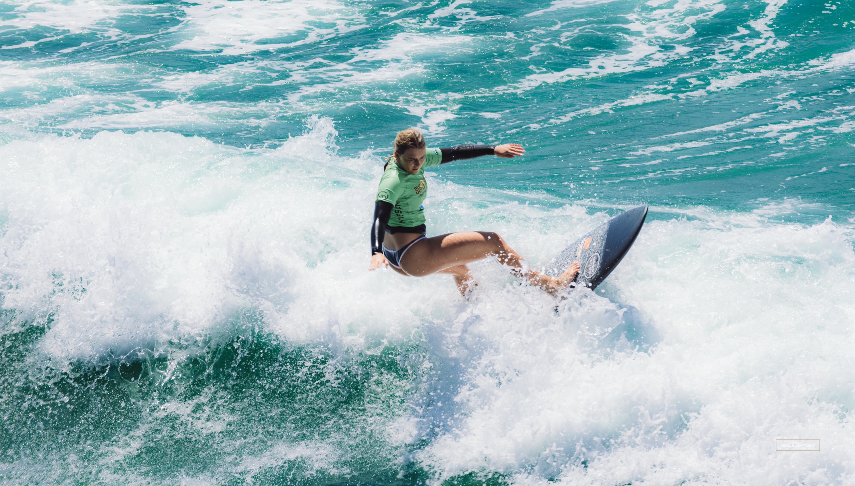 Dynamic action photograph of a young surfer carving through a breaking wave during the Super Girl Surf Festival at Oceanside Pier in Oceanside, California, photographed by Joe Gomez.